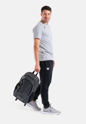 Zaino nero con una texture liscia e un logo, indossato da un uomo con maglietta grigio chiaro e pantaloni della tuta neri, con scarpe da ginnastica grigie.