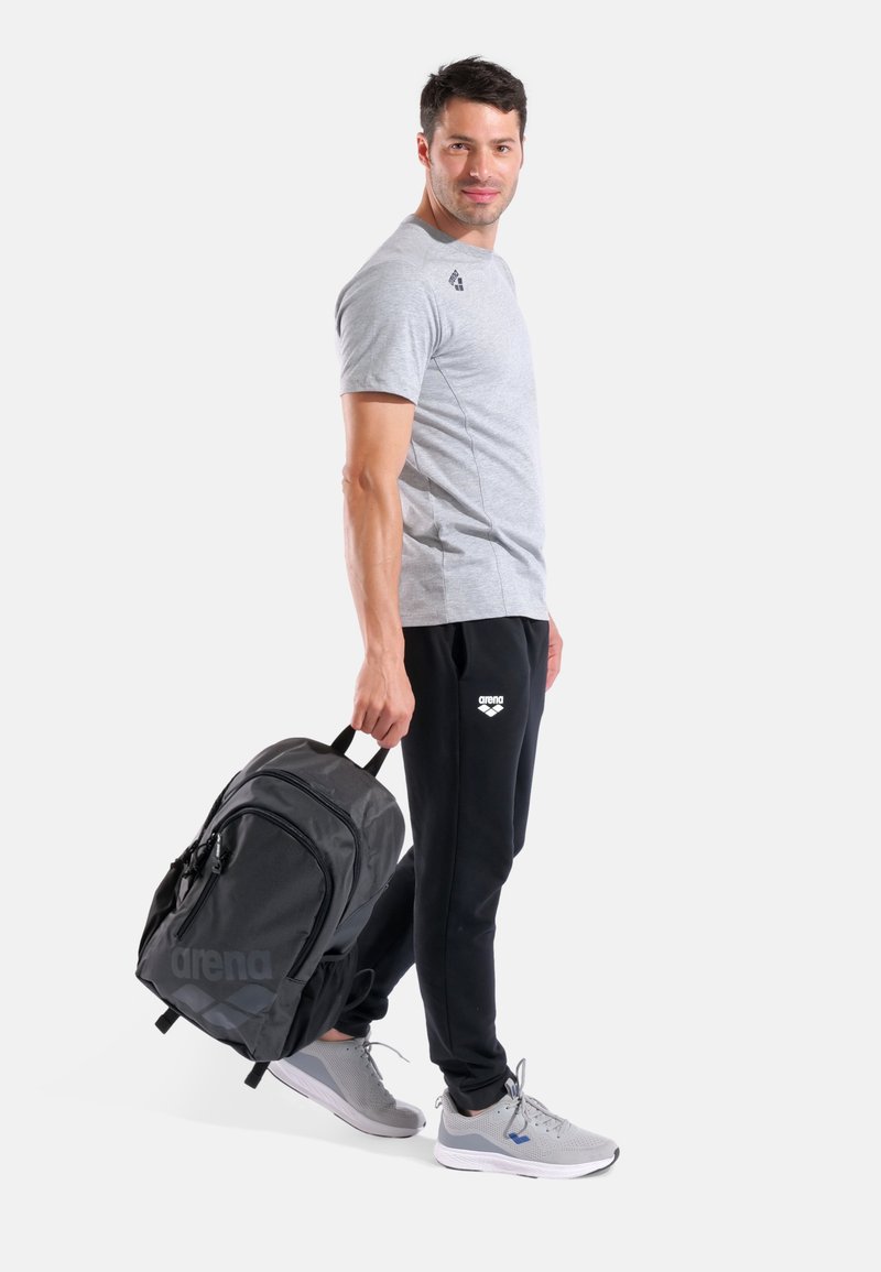 Zaino nero con una texture liscia e un logo, indossato da un uomo con maglietta grigio chiaro e pantaloni della tuta neri, con scarpe da ginnastica grigie.