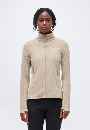 Žena oblečená v béžovej flísovej bunde so zipsom a otvorom na palce, čierne nohavice, stojaca na jednoduchom svetlom pozadí.