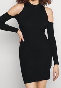 Robes en tricot noir avec un col haut, des épaules dénudées et des manches longues. Présente une silhouette ajustée et une texture côtelée.