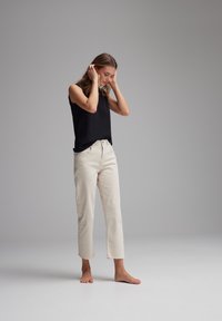 Top nero senza maniche abbinato a jeans cropped dritti beige chiaro. Modella in piedi a piedi nudi con le mani tra i capelli, sfondo minimale.