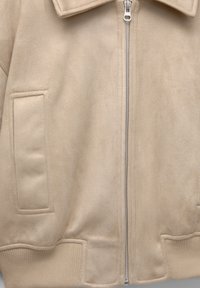Veste beige douce avec une fermeture éclair à l'avant, un col, deux poches carrées et des ourlets côtelés en bas et aux manches.