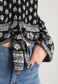 Blouse à motif floral noir et blanc nouée à la taille, associée à un jean en denim bleu, avec une main reposant dans une poche avant.
