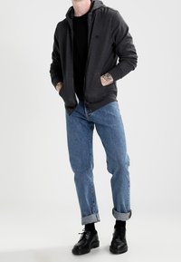 Un sweat à capuche noir zippé avec des poches avant, porté sur un t-shirt noir. Un jean bleu clair retroussé et des chaussures noires à lacets complètent l'ensemble.