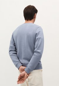 Lichtblauwe crewneck sweatshirt met lange sleeves, textuurstof en geribbelde manchetten, gedragen met lichtgekleurde broek, gezien vanuit de achterkant.
