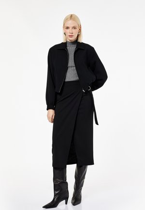 Frau mit kurzen blonden Haaren, die eine schwarze Jacke mit Reißverschluss, einen grauen gerippten Rollkragenpullover, einen schwarzen Wickelrock und schwarze kniehohe Stiefel trägt, steht vor weißem Hintergrund.