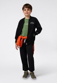 Svart bomberjakke, grønn t-skjorte, oransje hettegenser knyttet rundt livet, svarte joggebukser og grønne joggesko. Merkbare logo-detaljer synlige.