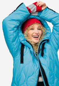 Light blue puffer jacket with a quilted pattern, black zippers, and adjustable drawstrings. Paired with a red knit beanie and striped gloves.