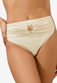 Hoogwaardige bikinionderbroek in crème, met een textuurpatroon van schubben, een geraffineerd ontwerp en een gouden schelpaccent in het midden.