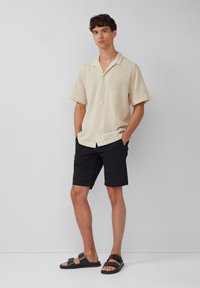Kortebloezem, beige, gestructureerde shirt met een knoopsluiting aan de voorkant, gecombineerd met zwarte shorts en zwarte sandalen, met banden en een platte zool.