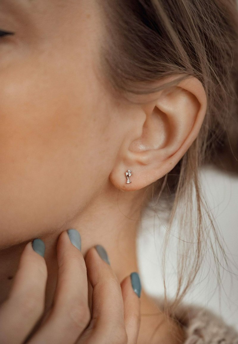 Orecchini a bottone in argento con un piccolo design floreale, caratterizzati da gemme trasparenti, appoggiati all'orecchio di un modello e su una pelle morbida e texturizzata.