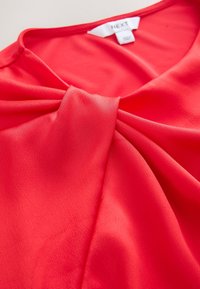 Blouse rouge corail vif avec un détail de nœud torsadé à l’encolure et une étiquette de marque "NEXT" visible à l’intérieur du col.
