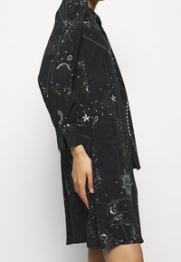 Černé šaty s hvězdným potiskem, strukturovaného střihu, s dlouhými rukávy, zapínáním na knoflíky na manžetách a jemnými záhyby na lemu pro zvýšenou pohybovost.