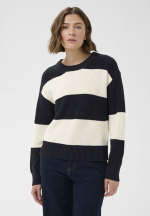 Žena s krátkymi hnedými vlasmi, oblečená v čierno-bielom pruhovanom pletenom svetri a tmavomodrých džínsoch, stojaca pred jednoduchým svetlým pozadím.