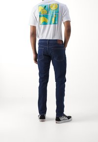 Blå denimjeans med smal passform, ihop med en vit t-shirt med en färgglad citrongrafik på ryggen. Svart och vit rutiga sneakers.