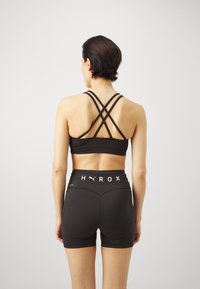 Crni sportski crop top s prečkama na leđima, u kombinaciji s crnim hlačicama visokog struka koje imaju bijeli "HYROX" logo.