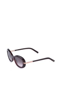 Lunettes de soleil ovales foncées avec branches noires et dorées, verres dégradés, posées en diagonale sur fond blanc.