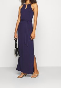 Mörkblå ärmlös maxikjol med nyckelhålsutskärning, bälte i midjan och sidoklyfta, tillsammans med metalliska sandaler och en svart handväska.