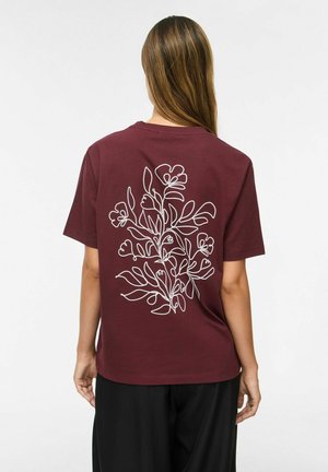Femme aux longs cheveux bruns porte un t-shirt bordeaux avec un dessin floral blanc en ligne au dos et un pantalon noir, debout sur fond blanc.