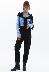 Modrá košile s dlouhým rukávem, černé kalhoty, černý svetr uvázaný kolem krku, hnědý klobouk a černé kotníkové boty na podpatku ve studiovém prostředí.