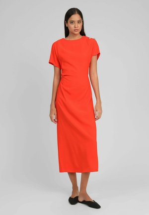 Femme debout portant une robe midi orange vif à manches courtes avec des boutons sur une épaule, assortie à des chaussures plates noires.