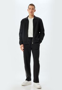Chaqueta negra de cremallera sobre una camisa blanca, combinada con pantalones de chándal negros que tienen rayas laterales blancas. El modelo lleva zapatillas blancas.
