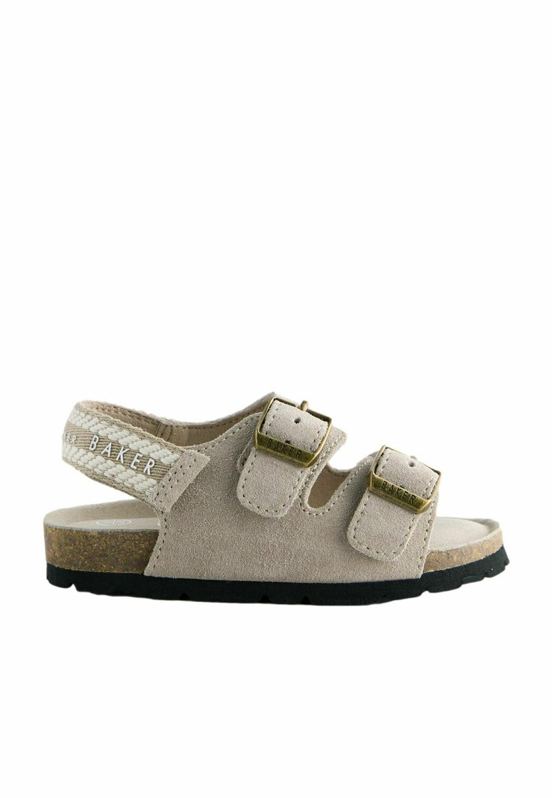 Beige mokkasandaali, jossa on kaksi säädettävää hihnaa ja kultasävyiset soljet. Korkkijalkapohja ja musta kumipohja, jossa on teksturoitu hihna.