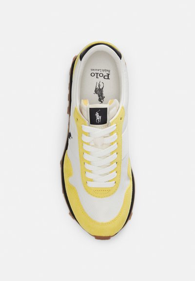 Ανδρικό αθλητικό παπούτσι με άνω μέρος από λευκό ύφασμα και κίτρινα σουέτ στοιχεία, στρογγυλεμένη μύτη, μαύρο λογότυπο και αντίθετη καφέ λαστιχένια σόλα.