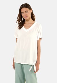 Top bianco a V in tessuto leggero, con maniche corte arrotolate e un dettaglio decorativo a nodo laterale all'orlo.