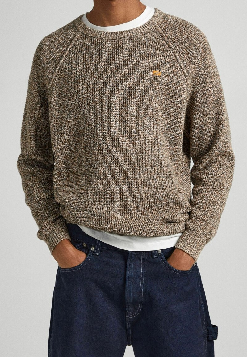 Maglione a maglia marrone con macchie, colletto a girocollo e polsini a coste, indossato sopra una T-shirt bianca, abbinato a jeans in denim blu scuro.