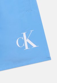 Ανοιχτό μπλε ύφασμα με λευκό λογότυπο "cK" τυπωμένο κοντά στο ραμμένο στρίφωμα, με λεπτομέρεια ραφής στο πάνω αριστερό μέρος.