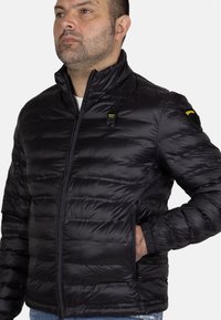 Mustat puff jacket, jossa on korkea kaulus, pystysuora tikkaustekstuuri, sivutaskut ja brändäys rinnassa ja olkapäässä. Suora pinta.