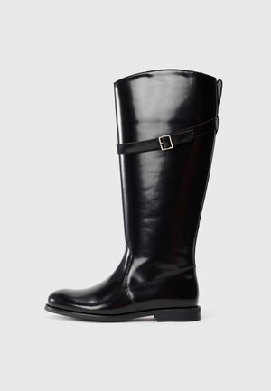 Botte en cuir noir haute avec un talon bas et une sangle décorative avec boucle en argent autour du haut du mollet.