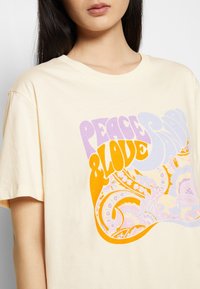 Mujer que lleva una camiseta de color beige claro con un colorido texto retro "Paz y Amor" y patrones florales abstractos en naranja, púrpura y azul claro.