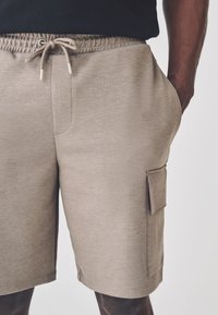 Pantaloncini beige in misto cotone con vita elastica, coulisse e tasca laterale. Tessuto liscio, lunghezza al ginocchio con vestibilità dritta.