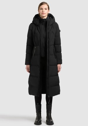 Femme portant un long manteau d'hiver noir matelassé avec une capuche, un pantalon noir et des bottines noires, debout devant un fond uni.