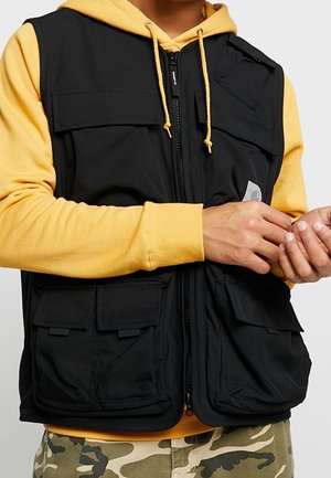 Personne portant un sweat à capuche jaune et un gilet noir multi-poches ajustant la manche, avec un pantalon camouflage visible en dessous.