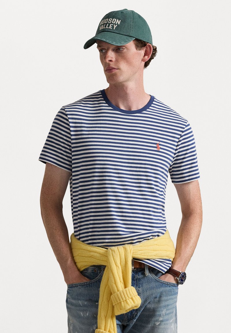 Rästripli t-särk siniste ja valgete horisontaalsete triipudega, ühtlane sinine krae, kantud kollase salliga talje ümber, aksessuaarina roheline müts.