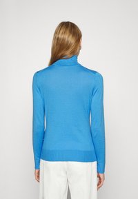 Pull à col roulé bleu à manches longues avec une texture lisse. Comprend un ourlet et des poignets côtelés. Porté avec un pantalon de couleur claire.