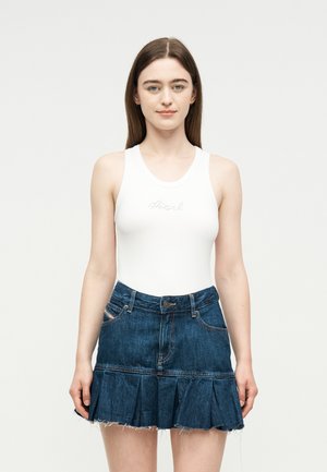 Noorpruunide pika juustega noor naine, kes kannab valget varrukateta tuba ja volditud tumel sinist teksaseelikut, seisab ühevärvilise tausta ees.