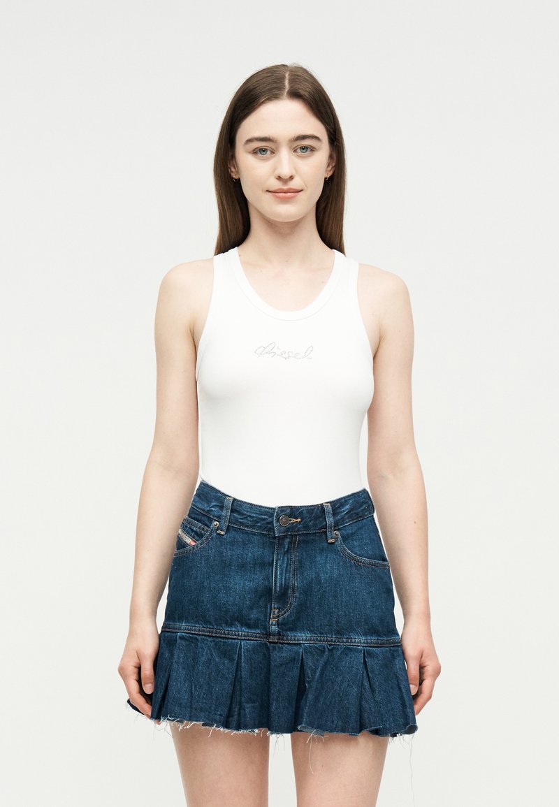 Νεαρή γυναίκα με μακριά καστανά μαλλιά, που φοράει λευκό αμάνικο τοπ και πλισέ σκούρο μπλε τζιν φούστα, στέκεται μπροστά σε απλή φόντο.