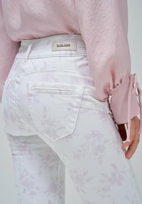 Jeans blancos con un estampado floral en morado claro, con bolsillos traseros. Combinados con una blusa rosa claro con mangas abullonadas.