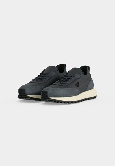 Paire de baskets à lacets noires et gris foncé avec semelles intermédiaires beiges et semelles extérieures noires texturées sur un fond clair uni.