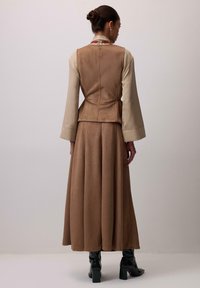 Barna velúr mellény világos bézs, hosszúkás ing fölött, amelyhez egy hasonló barna, légies szoknya és sima fekete magassarkú csizma társul.