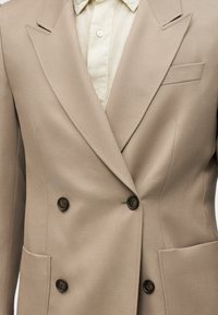 Blazer beige croisé avec quatre boutons foncés, larges revers, poches plaquées, porté par-dessus une chemise beige clair à boutons.