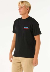 Sort bomulds T-shirt med et lille blå og rødt "RIP CURL" logo på venstre bryst, rund hals og korte ærmer, båret med beige bukser.