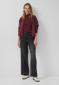 Vrouw die tegen een muur staat, gekleed in een bordeauxrode ritsjack, een bijpassend topje, donkere wijde jeans en witte sneakers.
