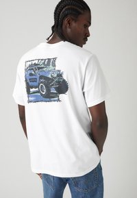 Balta trumpa rankovėmis marškinėliai su mėlynos off-road transporto priemonės grafika nugarinėje pusėje, turinčia tamsų kontūrą ir tekstą "Levi's".