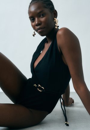 Fonott hajú nő, fekete ujjatlan ruhában, arany csattos övvel, tiszta világos háttér előtt ül.