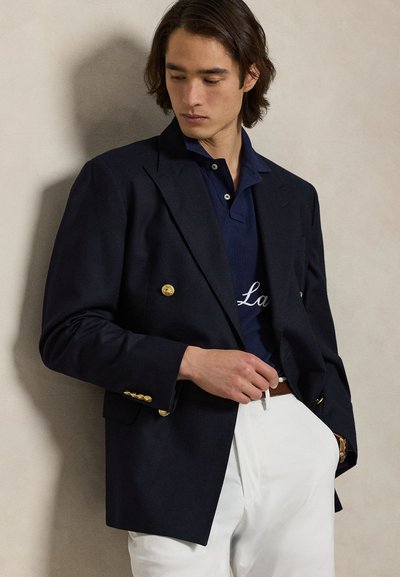 Tengerészkék dupla soros blézer arany gombokkal, tengerészkék galléros póló fehér hímzéssel alatta, fehér nadrággal és barna övvel kombinálva.